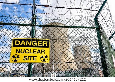 Close-up view of a yellow danger warning sign on the security fence with barbed wire of a nuclear power station with two cooling towers in a blurry background.
