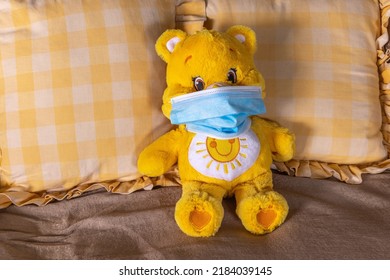 Close-up view of a yellow and white coloured teddy bear laying propped up by cushions on a sofa using a blue face mask during the Covid-19 pandemia. - Powered by Shutterstock