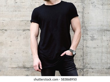 Closeup View Of Young Bearded Man Wearing Black Tshirt And Jeans Posing In Center Of Modern City. Empty Concrete Wall On The Background. Hotizontal Mockup