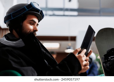 Close-up view of young man dressed in winter jacket browsing the internet on a digital device. Image showing male traveler with snowboarding gear engrossed in his tablet searching for snowy slopes. - Powered by Shutterstock