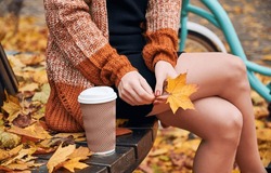Close-up View Of Young Woman With Coffee Is Sitting On Wooden Bench And Holding Yellow Leaf. Female Wearing Dress And Cagdigan Is Resting On Settle In Autumn Park. Concept Of Relaxation.