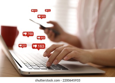 Closeup view of young woman using modern laptop with smartphone indoors and virtual dislike icons at table. Cyberbullying concept