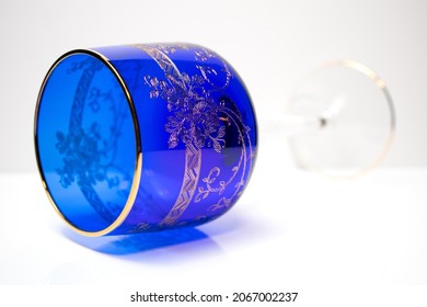 Closeup Of A Vintage Bohemian Crystal Wine Glass In Color Cobalt Blue Decorated With Gold On A White Background. Old Glassware Laying.