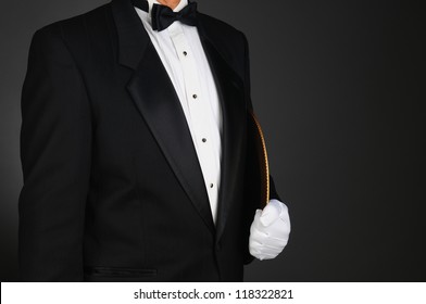 Closeup of a waiter in a tuxedo holding a serving tray under his arm. Horizontal format on a light to dark gray background. Man is unrecognizable.