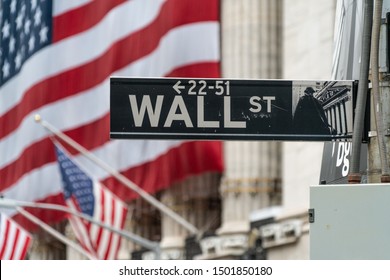 Closeup Of Wall St. Street Sign And Large New York City Financial Institution In Background With American Flag. The Most Economically Powerful City And The Leading Financial Center Of The World.