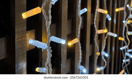 Close-up of warm white icicle lights strung against a wooden lattice backdrop. Water droplets cling to the bulbs, adding a touch of winter magic. Perfect for holiday and winter themes.