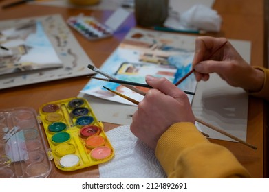 Closeup Of Watercolor Painting Process. Brush In Hand Above Painting. Colorful Medieval City Street On Paper Board.