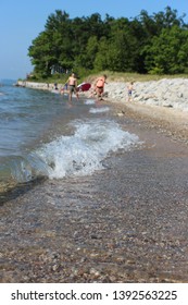 Closeup Of A Wave Rolling In Along A Roadside Beach Near Orchard Beach State Park On Lake Michigan, With A Family Playing Along The Beach In The Background.