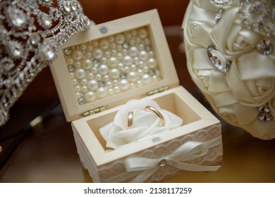 Close-up of the wedding rings of the bride and groom, in a white box, bridal tiara and white artificial bridal bouquet.