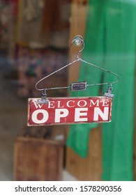 Close-up Of Welcome Open Sign Hanging From A Coat Hanger At The Entrance Glass Window / Door Of A Clothing Shop.