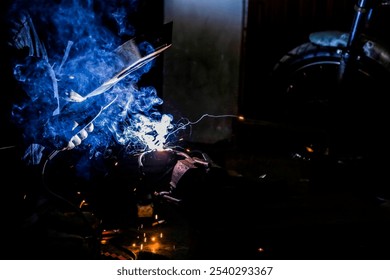 A close-up of a welder working on a motorcycle in a dark garage. The welder's protective gear and the bright welding torch are the only sources of light in the photo. The motorcycle is partially disas - Powered by Shutterstock