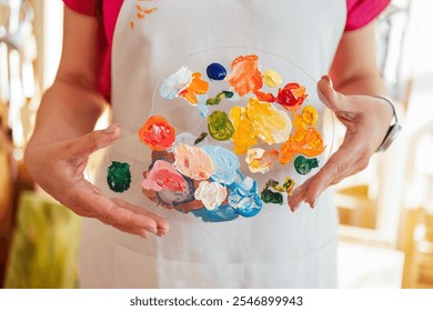 Close-up of well-groomed female hands holding plastic palette with mixed colors. Young woman in casual clothes and apron in art studio. Girl at painting workshop. Creative activity or hobby - Powered by Shutterstock