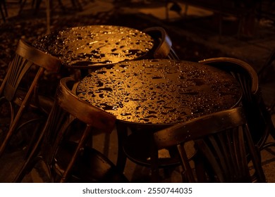 A close-up of wet cafe tables and wooden chairs, illuminated by warm, moody evening lighting. Raindrops shimmer on the tabletops, creating a serene and atmospheric scene. - Powered by Shutterstock