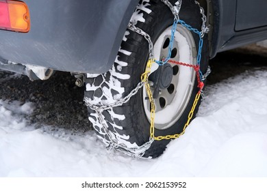 Close-up Wheel Of A Gray Car In Colored Iron Chains On The Snow, Winter Travel Safety Concept With Snow Chains On Tire