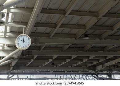 A close-up of a white clock hanging from the ceiling of a train station, with steel beams and infrastructure in the background.