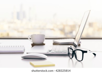 Closeup of white desktop with laptop, glasses, coffee cup, notepads and other items on blurry city background - Powered by Shutterstock