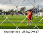 Close-up of a white diamond-patterned net in sharp focus, framing a blurred football field with players in action, creating a dynamic sports atmosphere.