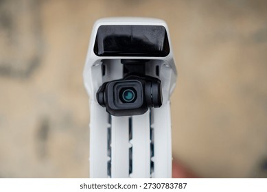 Close-up of a white drone's camera module with a black lens housing mounted on a textured surface showing the device's compact design for aerial photography or videography in outdoor environments.