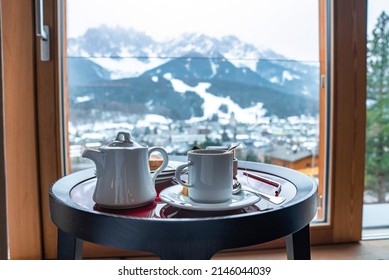 Close-up of white kettle and tea cup on table. Breakfast served with ceramics at resort. Mountains seen from glass window in background during winter.