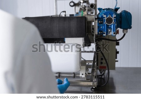 Close-up of a white plastic supplement container positioned on a semi automatic labeling machine inside a modern nutraceutical manufacturing facility. Industrial packaging process for whey protein and dietary supplements with hygienic production standards, automation technology, and pharmaceutical-grade equipment.