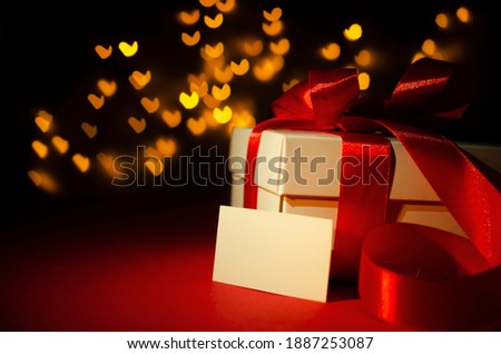 Closeup of white present box with red ribbon and bow, blank greeting card on the red surface against heart shape boken
