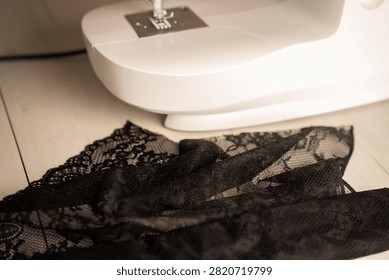 close-up of a white sewing machine base and metal presser foot with delicate black lace fabric draped across a light wooden tabletop; warm neutral tones, soft shallow depth-of-field, intimate mood