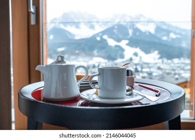 Close-up of white tea cup and kettle on stool. Breakfast served with ceramics on table at resort. Mountains seen from window in background during winter.
