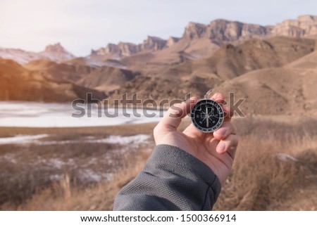Close-up wide angle male hand with a yellow watch bracelet holds a magnetic navigation compass against the backdrop of a beautiful landscape in the mountains with a winter mountain lake.