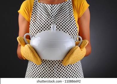 Closeup Of A Woman In An Apron And Oven Mitts Holding A Soup Tureen. Woman Is Unrecognizable, Horizontal Format On A Light To Dark Gray Background.