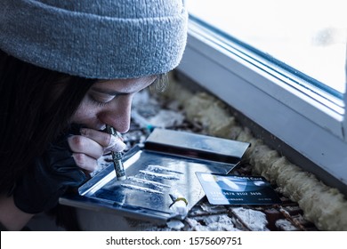Closeup woman in black fingerless gloves and hood, is inhaling white powder cocaine stripe. Addict teenage girl is taking coke, amphetamines in abandoned building. Drug addiction concept.