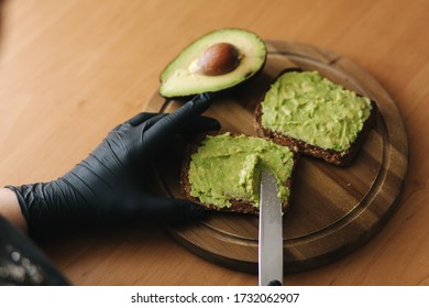 Close-up Of Woman In Black Glowes Puts Guacamole Or Avocado Spread On Top Of Rye Bread Toast On Wooden Board At Home. Vegan Breakfast