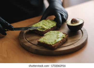 Close-up Of Woman In Black Glowes Puts Guacamole Or Avocado Spread On Top Of Rye Bread Toast On Wooden Board At Home. Vegan Breakfast