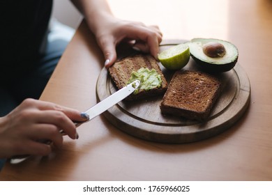 Close-up Of Woman In Black Glowes Puts Guacamole Or Avocado Spread On Top Of Rye Bread Toast On Wooden Board At Home. Vegan Breakfast