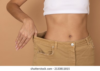 Closeup Of Woman Body In Large Pants With Hand Pulling Them To Show Slim Shape On Body Colored Background. Weight Loss By Keeping Diet And Exercising. 