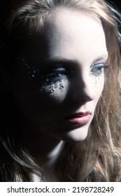 Close-up Woman Dark Portrait In Low Light Studio Shot. Model Face Partly Illuminated By Blue Light Imitating Television, Moon Light Or Nightlife. Model With Glued Shiny Jewelry Stones Around Eyes