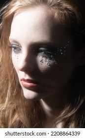 Close-up Woman Dark Portrait In Low Light Studio Shot. Model Face Partly Illuminated By Blue Light Imitating Television, Moon Light Or Nightlife. Model With Glued Shiny Jewelry Stones Around Eyes