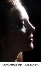 Close-up Woman Dark Portrait In Low Light Studio Shot. Model Face Partly Illuminated By Brown Light Imitating Television, Moon Light Or Nightlife. Model With Glued Shiny Jewelry Stones Around Eyes