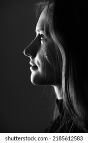 Close-up Woman Dark Profile Portrait In Low Light Studio Shot. Model Face Partly Illuminated By Light. Selective Focus And Image With Shallow Depth Of Field. Black And White Image