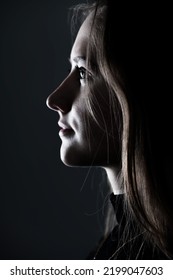 Close-up Woman Dark Profile Portrait In Low Light Studio Shot. Model Face Partly Illuminated By Light. Selective Focus And Image With Shallow Depth Of Field. Mysterious Atmosphere