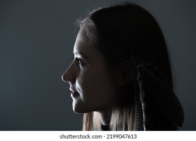 Close-up Woman Dark Profile Portrait In Low Light Studio Shot. Model Face Partly Illuminated By Light. Selective Focus And Image With Shallow Depth Of Field. Mysterious Atmosphere