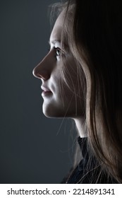 Close-up Woman Dark Profile Portrait In Low Light Studio Shot. Model Face Partly Illuminated By Light. Selective Focus And Image With Shallow Depth Of Field. Mysterious Atmosphere