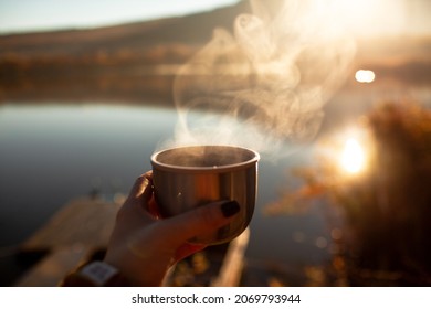 Close-up of woman hand holdin the cup of tea with stamp against the sun and lake in the nature