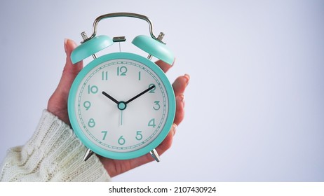 Close-up of woman hand holding blue retro alarm clock white background,time on clock is ten o'clock ten minutes,10:10.Time to wake up,early morning.Blue monday day concept.Copy space.Turquoise clock
