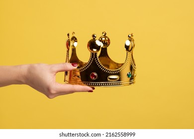 Closeup of woman hand holding golden crown, concept of awards ceremony, privileged status, superior position. Indoor studio shot isolated on yellow background.