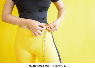 Close-up of woman hands is measuring waistline using yellow measure tape isolated on bright yellow background. Copy space for dietary and healthy lifestyle concept.