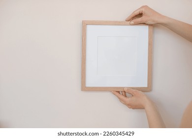 Close-up of woman hanging square wooden frame with empty mockup copy space on wall. Minimal art concept. - Shutterstock ID 2260345429