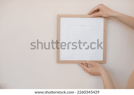 Close-up of woman hanging square wooden frame with empty mockup copy space on wall. Minimal art concept.