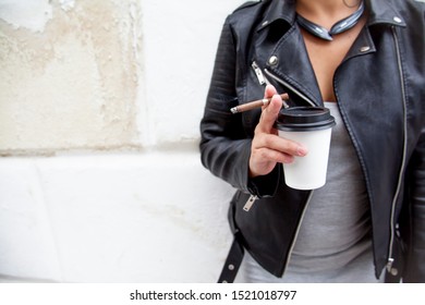 Close-up Of The Woman Holding Cigarette And Coffee Background City Wall. Wearing Grey Dress, Headphones And Black Jacket.