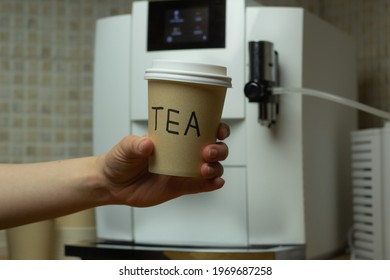 Closeup Of A Woman Holding A Disposable Tea Cup With Coffee Machine In The Background.