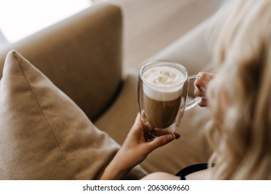 Closeup of a woman holding a double glass cup of coffee with foam, sitting on sofa at home.